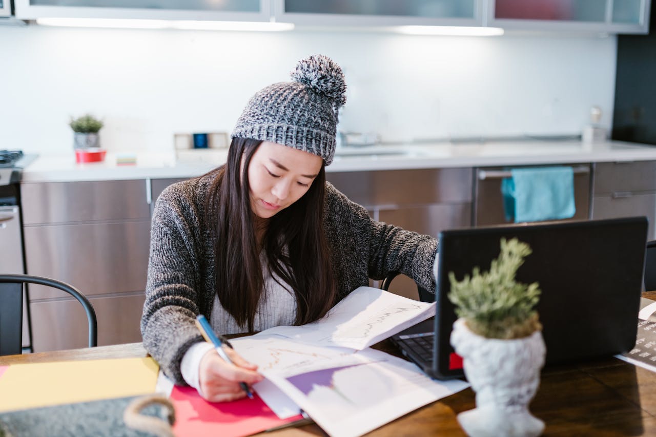 Asian woman in cozy attire working on documents with a laptop at home office desk.